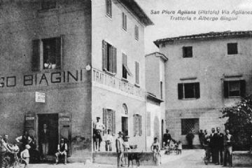 La cartolina fu commissionata ai primi del ‘900 dai proprietari dell’albergo trattoria Biagini situato nel palazzetto Melani; accanto, tramite un portone ad arco si accedeva alla piccola corte. Davanti si vede il palazzetto Masi che in quest’epoca occupato dalla caserma dei Regi Carabinieri. In questo edificio trovarono alloggio per qualche anno le suore Mantellate venute da Treppio nel 1876 per fondare la seconda casa di questa congregazione. La caserma fu trasferita a metà degli anni Settanta e il palazzo fu demolito per realizzare il nuovo cinema di Nocino, che a sua volta, a seguito di un incendio non venne riaperto e le sue rovine furono abbattute per realizzare l’attuale galleria.