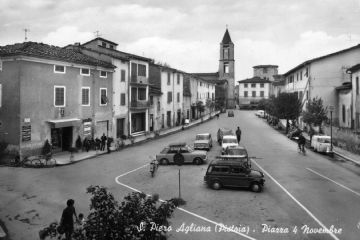 Piazza IV Novembre, anni ‘60. La cartolina mostra una situazione in cambiamento: tra il 1950 e il 1960 fu ristrutturato il campanile su disegno degli architetti fiorentini Sante e Savioli.<br>Nella piazza compaiono le macchine parcheggiate in quella che diverrà un’ingombrante aiuola spartitraffico togliendo respiro a questo spazio pubblico oggi destinato a parcheggio.