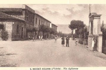 San Niccolò Agliana – Via Aglianese. Questa cartolina realizzata intorno al 1940 mostra in primo piano, sulla destra, il grande tabernacolo realizzato ai primi del XIX secolo, demolito durante gli anni ‘50/60 per essere ricollocato in una posizione più arretrata a seguito dell’allargamento della strada. Dal lato opposto della via si notano alcuni edifici che sembrerebbero di recente costruzione, ma, in verità, il secondo edificio partendo da sinistra fu edificato durante la metà del 1800 per accogliere il Teatro dell’Unione.